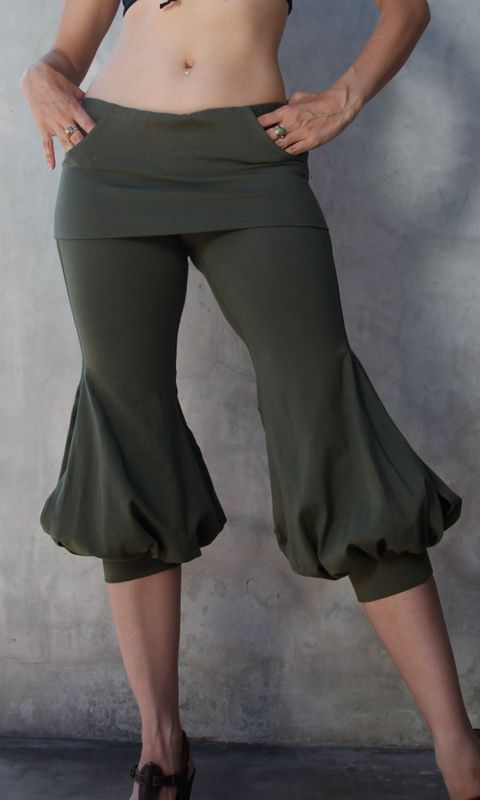 a woman is standing and posing, wearing a pair of green  pants with a short skirt layered over them. she has brown shoes on.  the background is a grey wall.