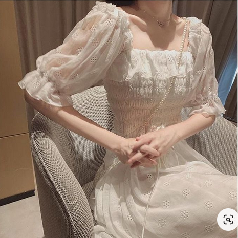 a woman wearing a flowy white summer dress is posing in a seated position, her hands clasped in front of her. the dress features delicate lace details, puff sleeves, a square neckline, and a ruched bodice. the soft, natural lighting creates a romantic and feminine atmosphere.