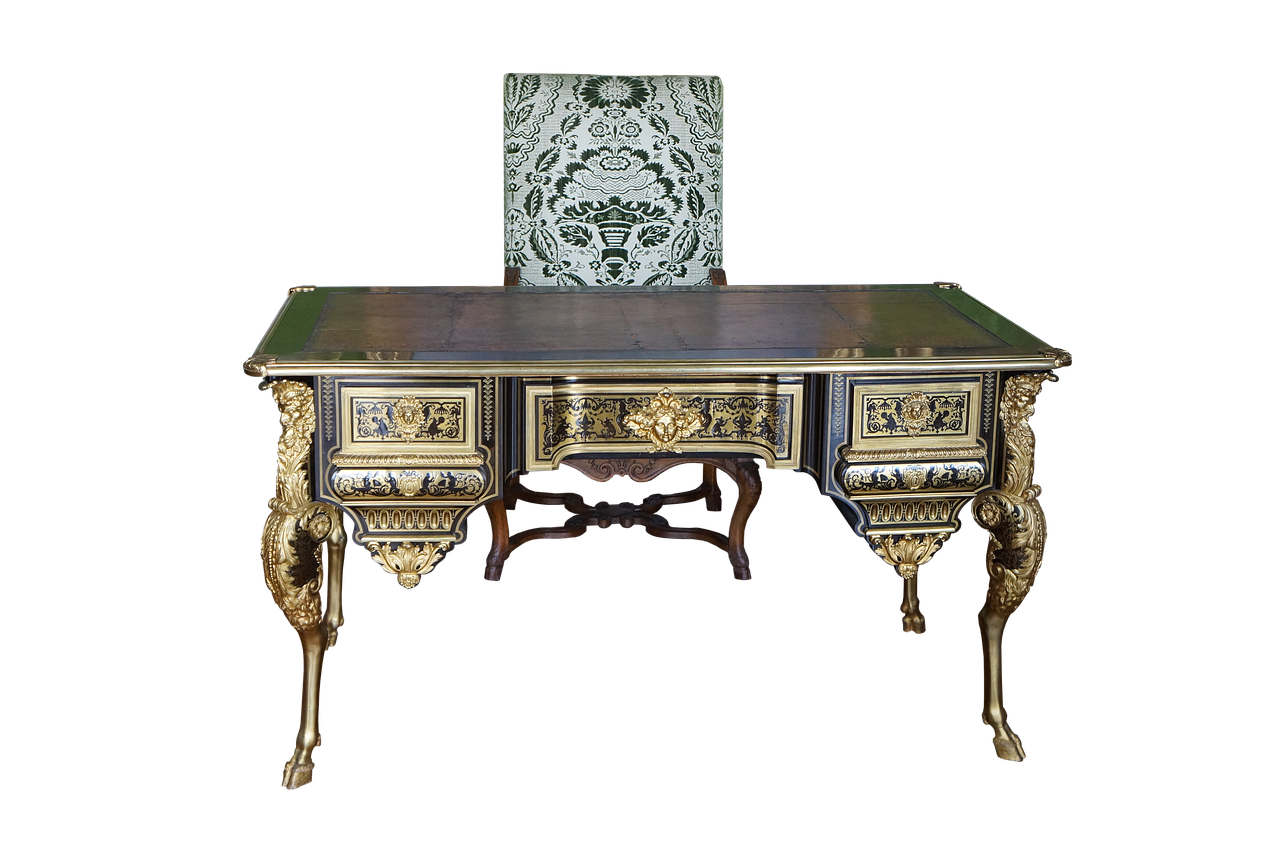 the image shows an antique desk and chair set against a black background. the desk is elaborately decorated with gold accents and carvings. it has several drawers with intricate designs. the legs are curved and ornate. the chair is upholstered with a green and white patterned fabric.