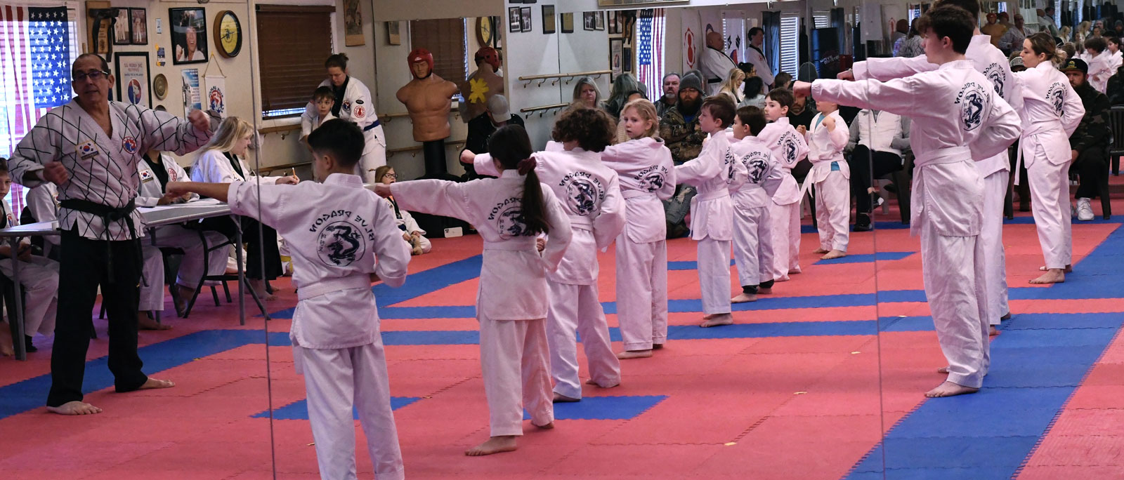 a group of people are practicing martial arts in a studio. there are both kids and adults, wearing white uniforms, belts, and black belts. they are practicing punches and kicks. the studio has blue and red floor mats.  the walls are lined with mirrors, which reflect the people in the studio. there is a large group of children in white uniforms, practicing their stances and moves. the children are learning from an instructor and are paying close attention to the instructor’s instructions. the instructor is demonstrating the proper technique, and the children are trying their best to copy him.  the image is a great example of the benefits of martial arts training. it’s a great way to get exercise, learn discipline, and build selfconfidence.