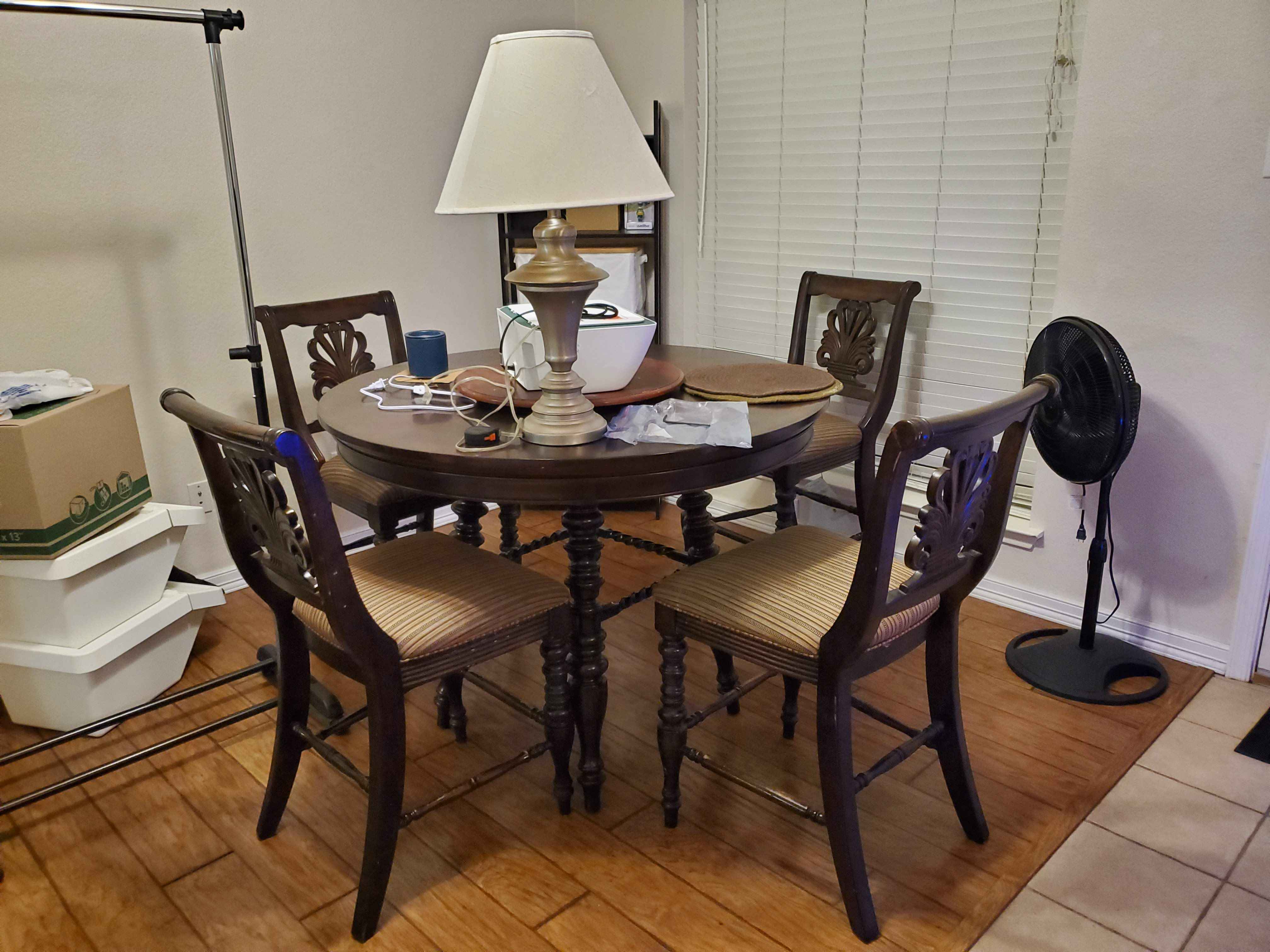 this image contains a round, dark wood table with four chairs surrounding it. a lamp sits on the table, with several objects scattered around it. behind the table, there is a window with white blinds. a black fan stands to the right of the table. to the left of the table, there are boxes stacked on top of each other and a coat rack.