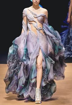 a woman in a beautiful gown with blue and purple patterns is walking the runway, she is wearing a high heel silver shoes. she is looking elegant and glamorous. the gown has a unique design and the fabric is flowing and light. the dress is an example of haute couture fashion.