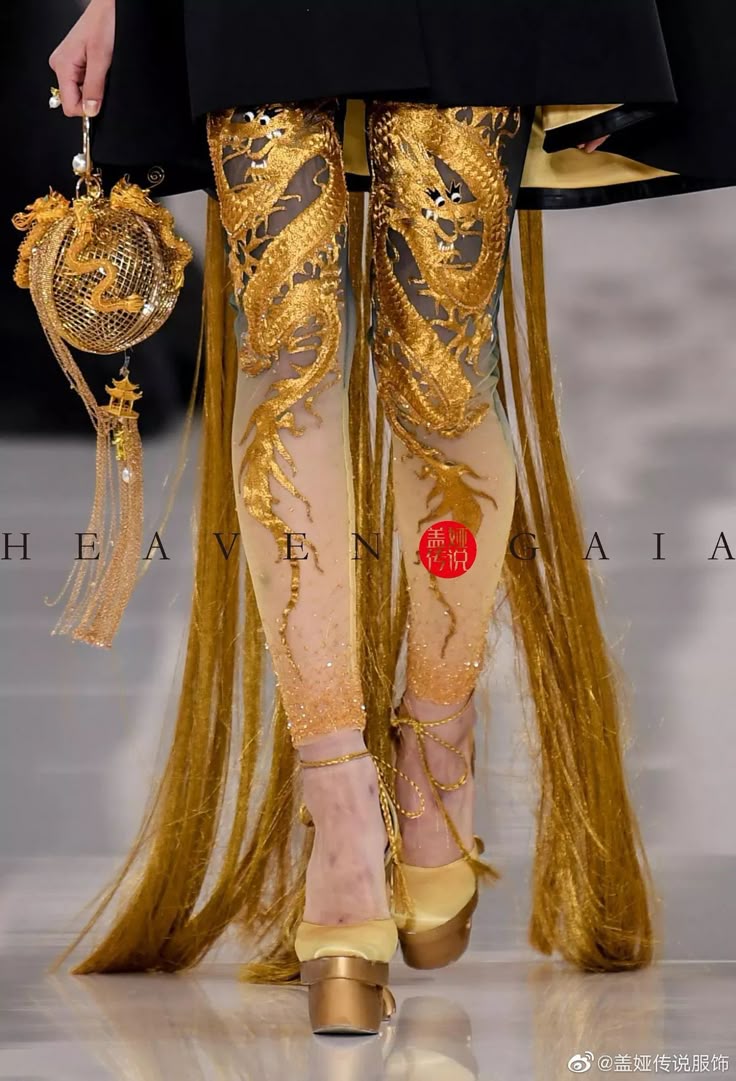 closeup of a model on a runway showcasing a heaven gaia design. the model is wearing sheer tights with elaborate gold dragon embroidery on the legs, paired with gold platform heels that lace up the ankles. she's holding a spherical gold handbag with a tassel detail. the outfit is complemented by long strands of gold fabric cascading down, and the backdrop is a lightcolored runway surface.