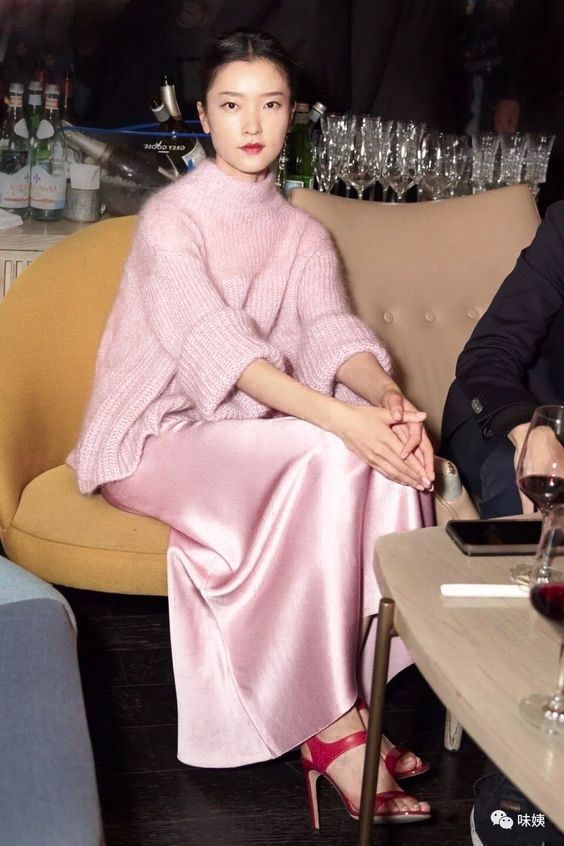 a young asian woman, possibly chinese, is elegantly seated in a yellow chair in what seems to be a bar or restaurant setting.  she's wearing a pale pink oversized knit sweater and a matching silk slip skirt.  she has on stylish red highheeled sandals.  the background features wine bottles, wine glasses, and a partially visible person in a dark suit.  the overall ambiance is sophisticated and nighttime. the lighting is dim and atmospheric. the image appears to be a professionally taken photograph, possibly for a fashion publication or event.