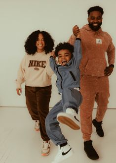 a happy african american family of three is captured in midair, all smiles as they enjoy a playful moment together in a studio setting. the father, on the right, lifts their son in the air as he beams with joy, while the mother, on the left, holds their son's other hand and laughs. they are all stylishly dressed in athleisure wear. the minimalist white background emphasizes the family's joyful connection and the soft, natural lighting enhances the heartwarming mood.