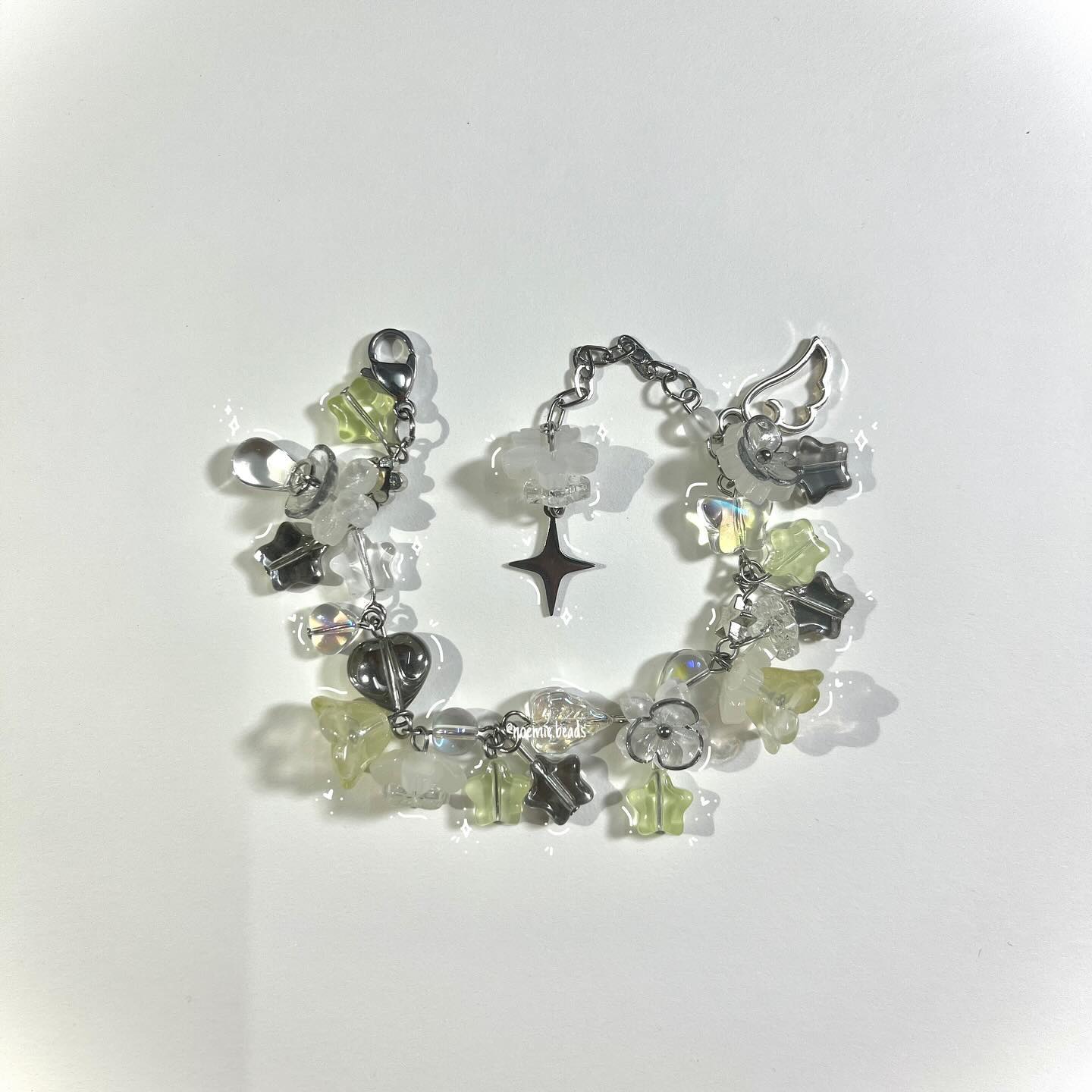 a bracelet made of silvercolored metal with clear, white, and pale green translucent beads and charms in the shapes of flowers, stars, hearts, and a cross. an angel wings charm is also visible. a clasp is visible.