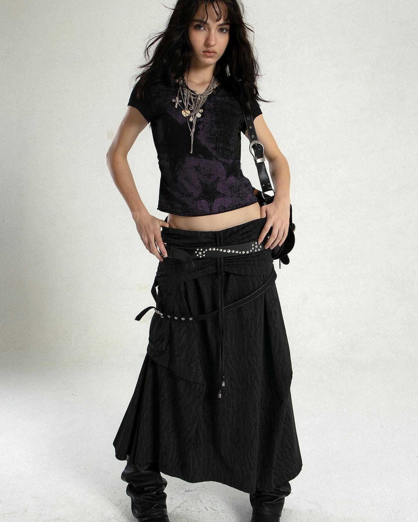 a fullbody shot of a young woman with long dark hair, wearing a dark purple textured tshirt, a long black asymmetrical skirt with a studded belt, and black boots. she is accessorized with a silver necklace and a black shoulder bag. the background is plain white.  she appears to be modeling the outfit. the overall style is dark, edgy, and fashionable.
