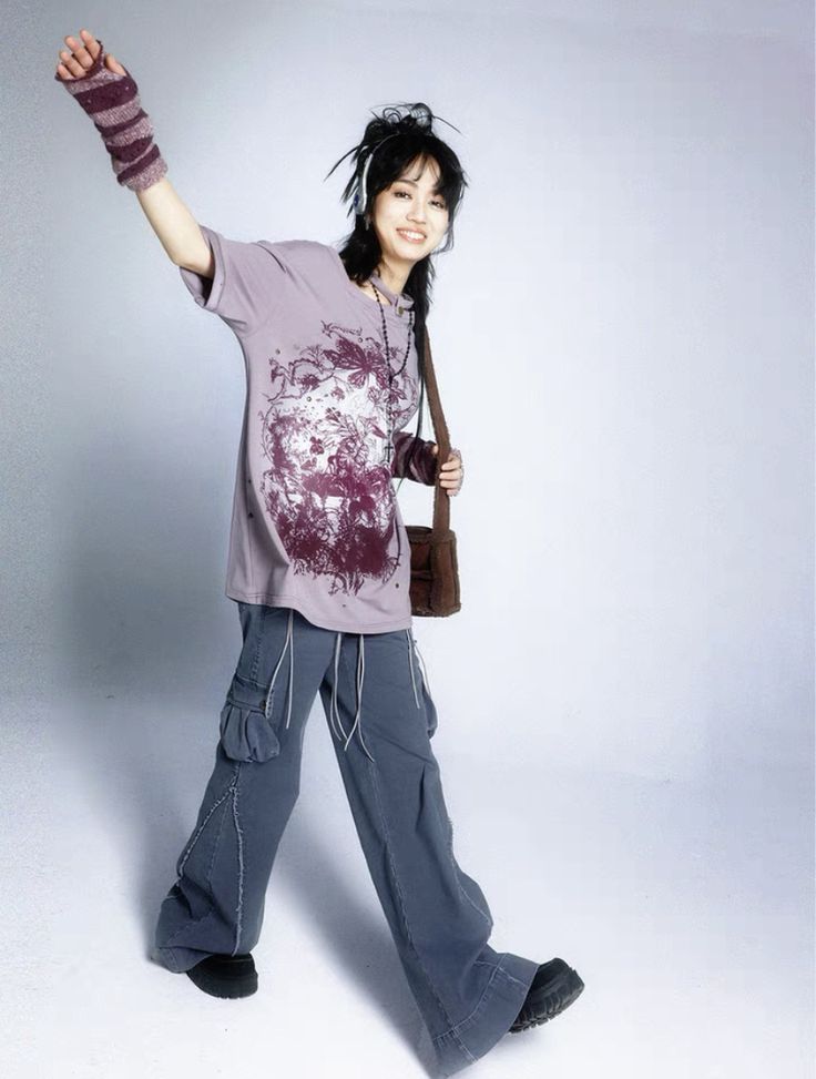 a young asian woman is depicted in a fullbody studio shot against a light gray background. she's showcasing a casual outfit consisting of an oversized, light purple graphic tshirt with a dark print,  wideleg gray cargo pants, black chunky boots, and fingerless gloves. she also carries a brown crossbody bag. her pose is dynamic and expressive, suggesting a streetstyle or grunge aesthetic with hints of 90s and y2k influences. the overall feel is relaxed and fashionable.