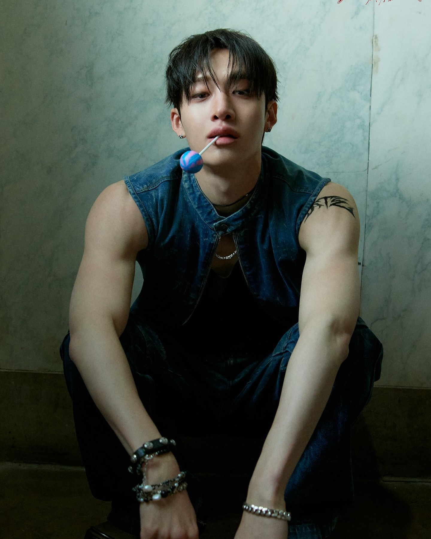 the image is a closeup portrait of a young asian man, possibly a kpop idol, who is sitting with a lollipop in his mouth. he's wearing a denim vest over a dark shirt and jeans.  he has dark hair, noticeable tattoos on his arm, and several bracelets adorning his wrists. the lighting is moody and dark, creating a dramatic and stylish effect. his expression is intense and serious.  the overall aesthetic is edgy and fashionable.