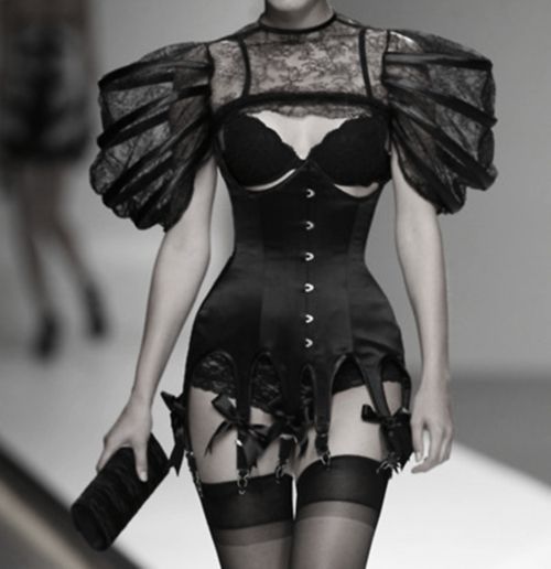 a black and white image shows a model on a runway wearing a black couture lingerie dress featuring a corset, puff sleeves, lace detailing, garters and stockings. she is carrying a clutch purse.