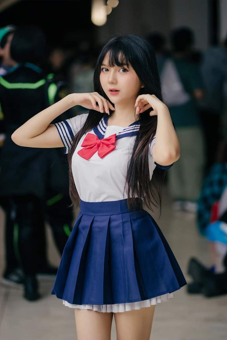 a young asian woman is posing for a photo while wearing a sailor uniform. the uniform consists of a white top with blue striped trim and a red bow, paired with a blue pleated skirt. she has long black hair and is standing indoors, possibly at a convention. the background is blurred with other people in the scene.