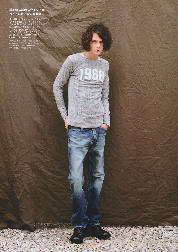 a full shot of a male model standing with hands in pockets, wearing a grey longsleeved sweater with "1968" printed on it, paired with blue denim jeans and black boots, against a brown backdrop. the ground is covered in pebbles. there is japanese text visible in the top left corner.