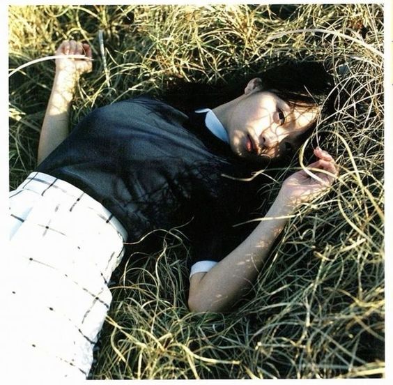 a young woman in a black and white outfit is lying on her side in a field of tall, dry grass. she is looking at the camera with a neutral expression. the photo is taken from above, giving an aerial perspective. the lighting is natural and soft. the overall color palette is muted and earthy.