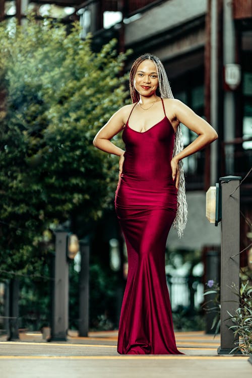 the image shows a smiling young woman standing confidently on a walkway. she's wearing a long, elegant, burgundy silk or satin dress that flows to the floor. the dress has a simple, formfitting design and a low vneck. she has long braids with gray and black strands. the background includes greenery, trees, and a building, giving a natural and sophisticated ambiance. the overall impression is one of beauty, elegance, and confidence.