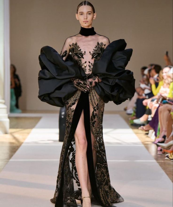 a model walks down the runway wearing a stunning black gown with gold detailing and a dramatic, puffedup sleeve. the dress is made of delicate lace and features a thighhigh slit that showcases the model's legs.  the model is wearing minimal makeup and jewelry, letting the dress take center stage. the runway is lined with seated spectators, all captivated by the show.