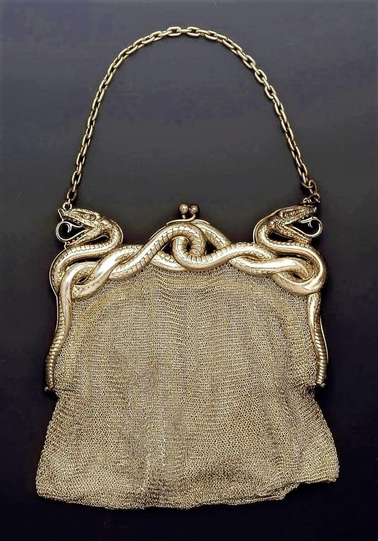 the image shows an antique metal mesh purse with a distinctive snake design. the frame of the purse is adorned with two coiled silver snakes facing opposite directions with their mouths slightly open. the heads of the snakes connect to a chain handle. the purse is constructed from a finely woven metal mesh, giving it a metallic sheen. the clasp of the purse is located in the center. the background is a plain dark color that makes the purse stand out.