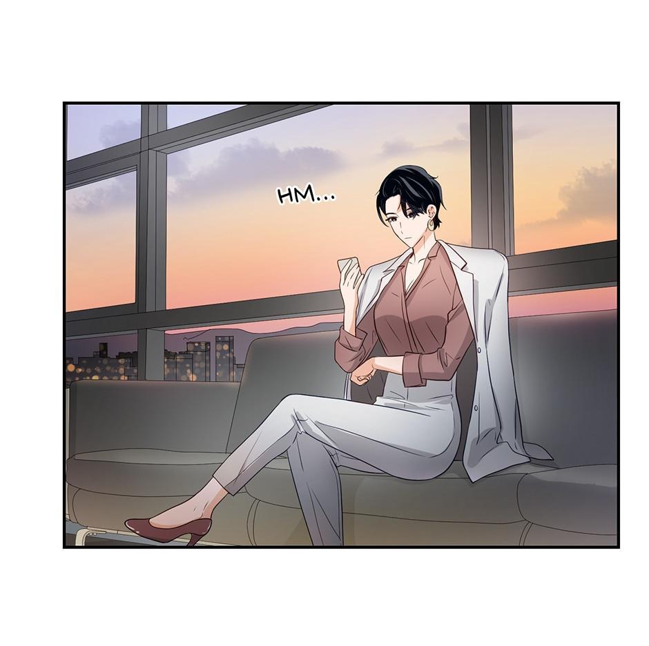 the image shows a stylish woman sitting on a couch in a modern office.  she is elegantly dressed in a pantsuit and blazer, and is holding a phone while looking thoughtful. the setting sun casts a warm glow through the large window behind her, offering a view of a cityscape. the overall mood is calm and sophisticated.