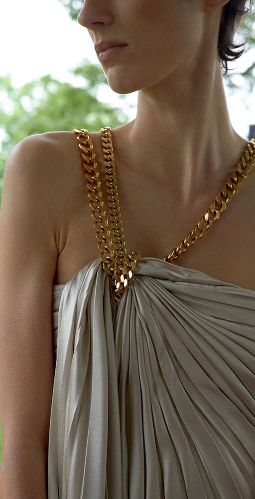 a woman is shown in a closeup shot from the upper torso up. she is wearing a dress with pleated, lightcolored fabric that drapes from the shoulder. the dress is held up by thick, goldcolored chain straps that go over each shoulder. the straps appear to be intertwined at the top of the dress, creating a focal point. the woman is shown in profile, revealing her neck and shoulder. her skin is light, and she has short, dark hair. in the background, there is a glimpse of green foliage, suggesting an outdoor setting.