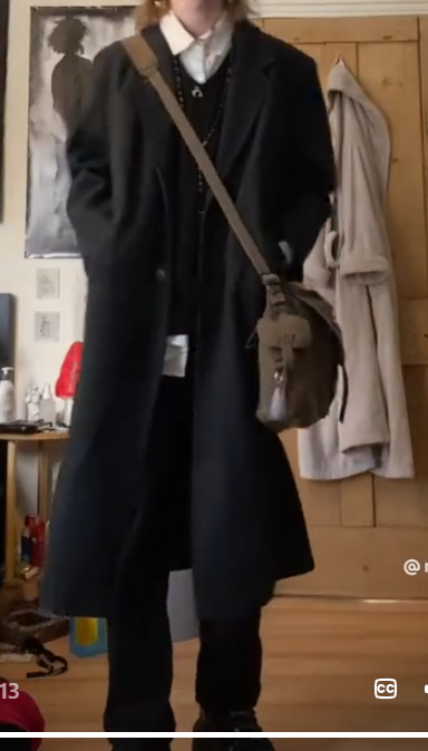 a young woman in a black coat stands in a room with wood flooring. she is wearing a black shirt, a white collared shirt peeking out from underneath, black pants, and black boots. she also has a black crossbody bag with a fringe detail and a silver chain necklace with a black pendant. the woman is wearing a black beret. there is a white bathrobe on the wooden door behind her, and a drawing on the wall next to her. the room is decorated in a minimalist style.