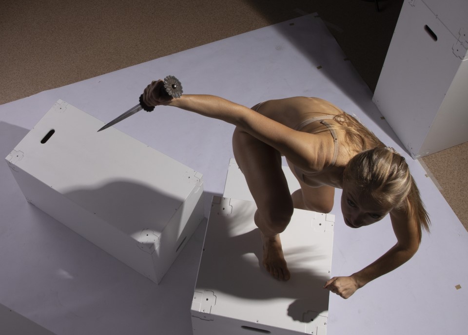 an overhead studio shot shows a slender fairskinned woman with long blonde hair wielding a knife while standing on top of several white boxes. she wears a lightcolored bra and is barefoot. her body is bent forward, with one arm outstretched holding the knife and the other arm bent down towards another box. she is looking down with a focused expression.