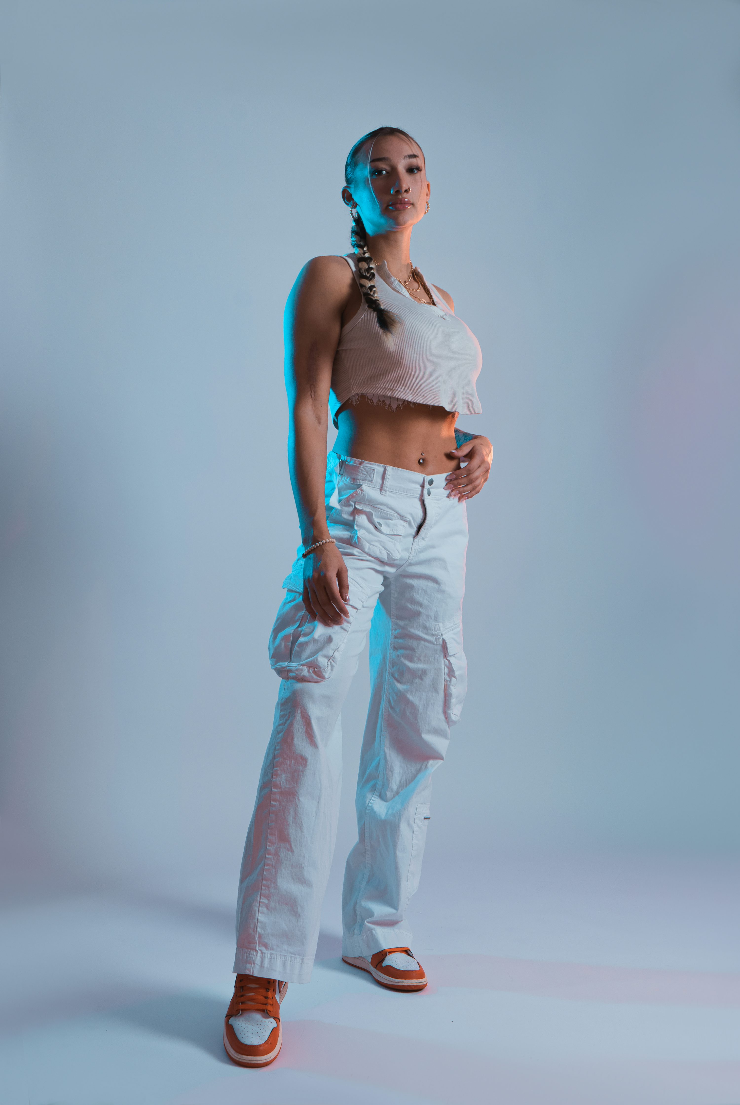 the image showcases a young, fit woman confidently posing against a light blue studio background. she's dressed in stylish streetwear: a white cropped tank top, loosefitting white cargo pants, and orange sneakers. her hair is styled in braids.  the lighting is dramatic, with a cool blue hue accentuating her pose. the overall aesthetic is fashionable and modern.