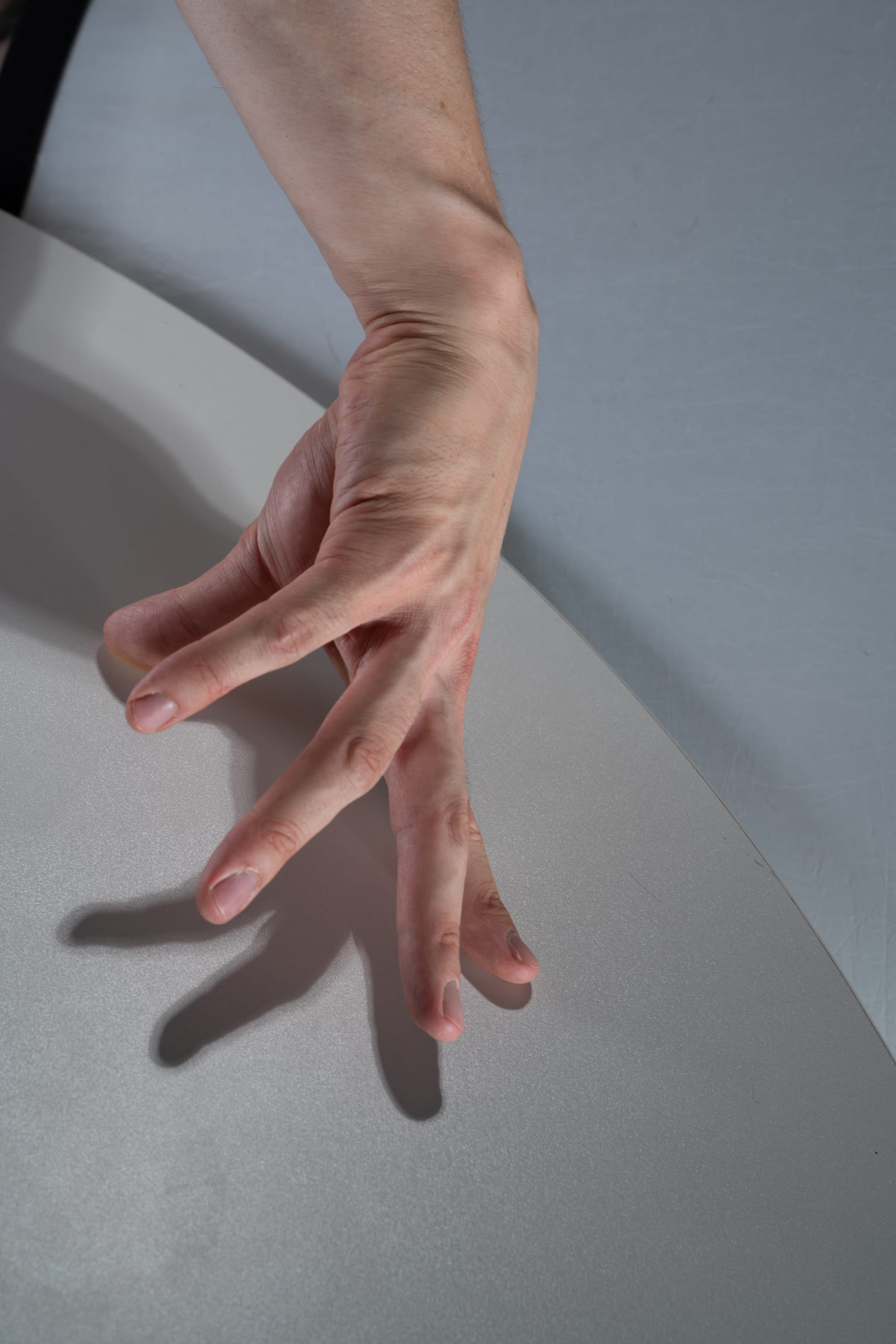 a closeup shot of a hand with long thin fingers spread out and casting a shadow on a white surface. the arm is extended and the hand is relaxed, with the fingers slightly curved. the lighting is soft and even, highlighting the details of the hand and the shadow it casts. the background is a neutral gray, which helps to focus attention on the hand.