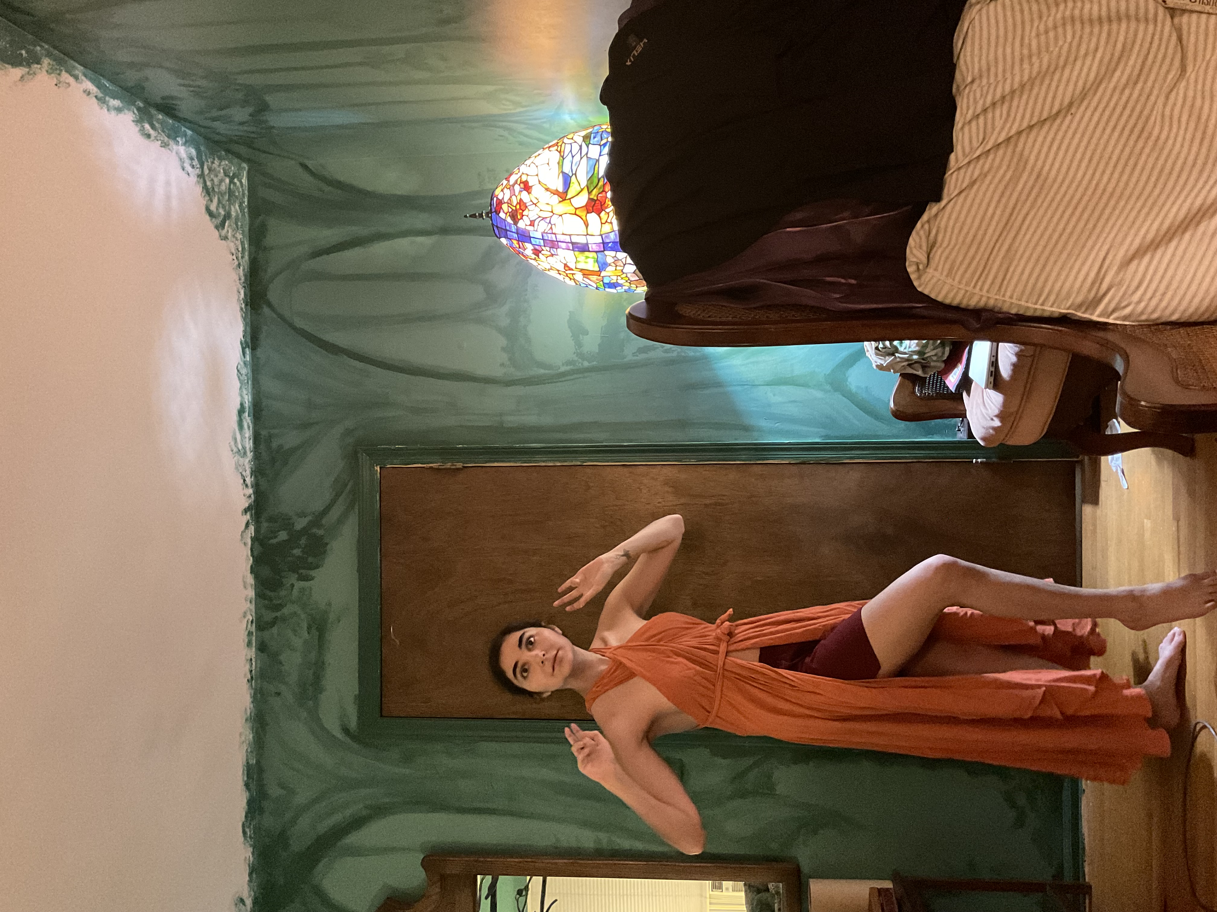 a young woman is standing in a room with green walls that have a painted design resembling trees. she is wearing a long, orange halter dress that is wrapped and tied at the waist.  the dress has a high slit on one side, showing her legs. she is posing with her hands raised. in the background, there's a stainedglass lamp on a wooden chair and a wooden door. the room appears to have a vintage or retro feel.  the overall atmosphere is artistic and somewhat bohemian.