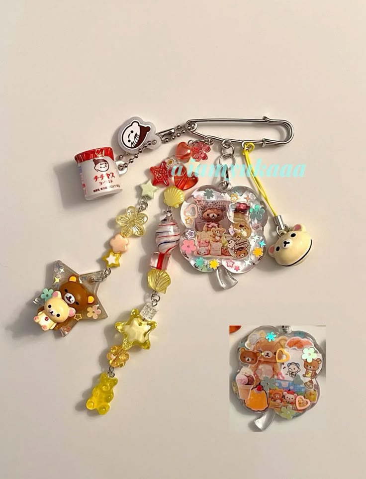 the image shows a charm attached to a safety pin. the charm consists of several smaller trinkets, including rilakkuma bear figures, stars, colorful beads, cartoon character miniatures, and sweetsthemed decorations.