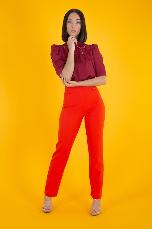 a fulllength studio shot of a woman wearing red pants and a maroon blouse, standing against a bright yellow background. she is posing with her hand on her chin, looking directly at the camera. she is wearing heels and has short hair. the image showcases fashion and portrait photography.