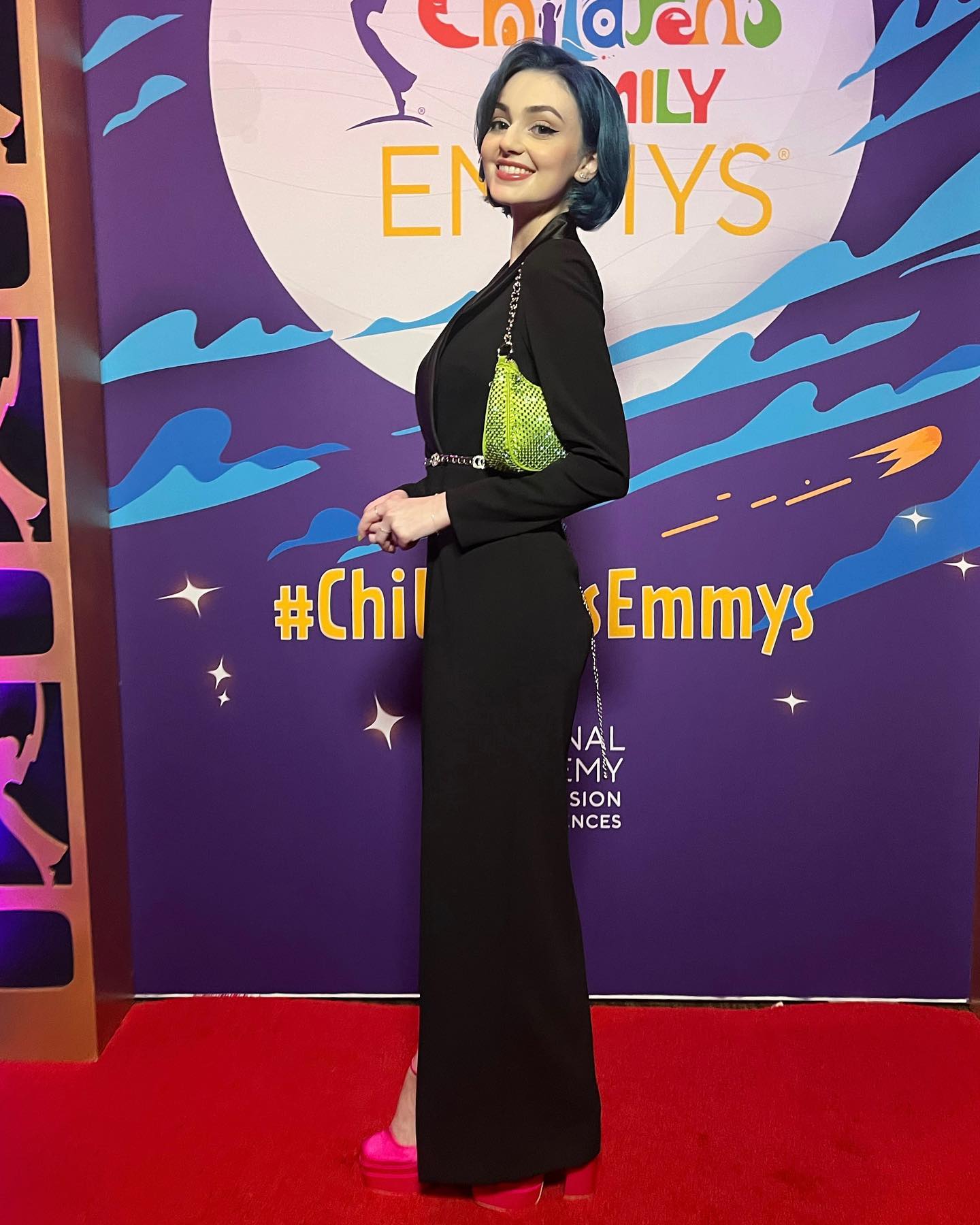 the image shows a woman on a red carpet at a children's emmys awards ceremony. she is wearing a stylish black jumpsuit with a side slit, vibrant pink heels, and a bright lime green purse. her hair is a striking blue. the background is a purple backdrop with the children's emmy awards logo. the overall impression is one of a formal event and fashionable attire.