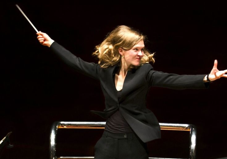 a female conductor stands on a dark stage with a spotlight shining on her. she is dressed in a black suit and is conducting an orchestra with a baton. her hair is long and blonde, and she is smiling as she directs the music.
