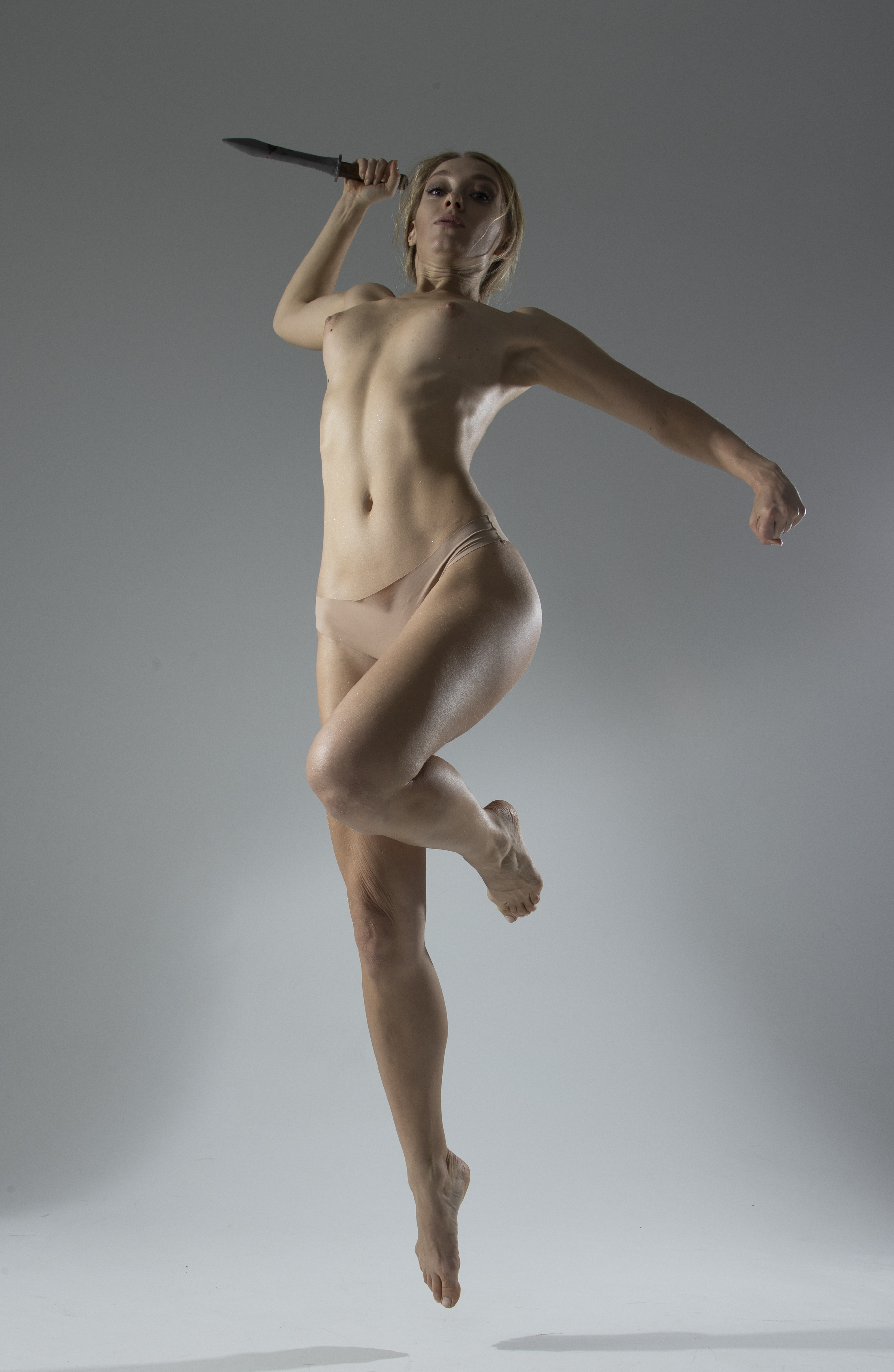 a nude woman is captured in a dynamic pose midair, holding a sword as if preparing to throw it. her body is athletic and toned, and she is slender. the lighting in the studio shot is dramatic, highlighting her form against the light background. her pose suggests a warrior in action, fighting or preparing for battle.