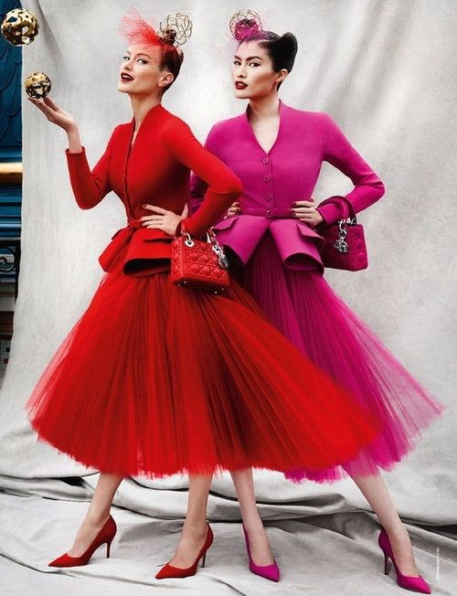 two female models are posing for a fashion shoot. the model on the left is wearing a red dress, and the model on the right is wearing a pink dress. both dresses are made of tulle, which gives them a voluminous and dramatic look. both women are wearing high heels. the model on the left is holding a small red handbag, while the model on the right is carrying a larger black handbag. both are wearing hats on their heads. the background is a plain white wall. they look elegant and glamorous.