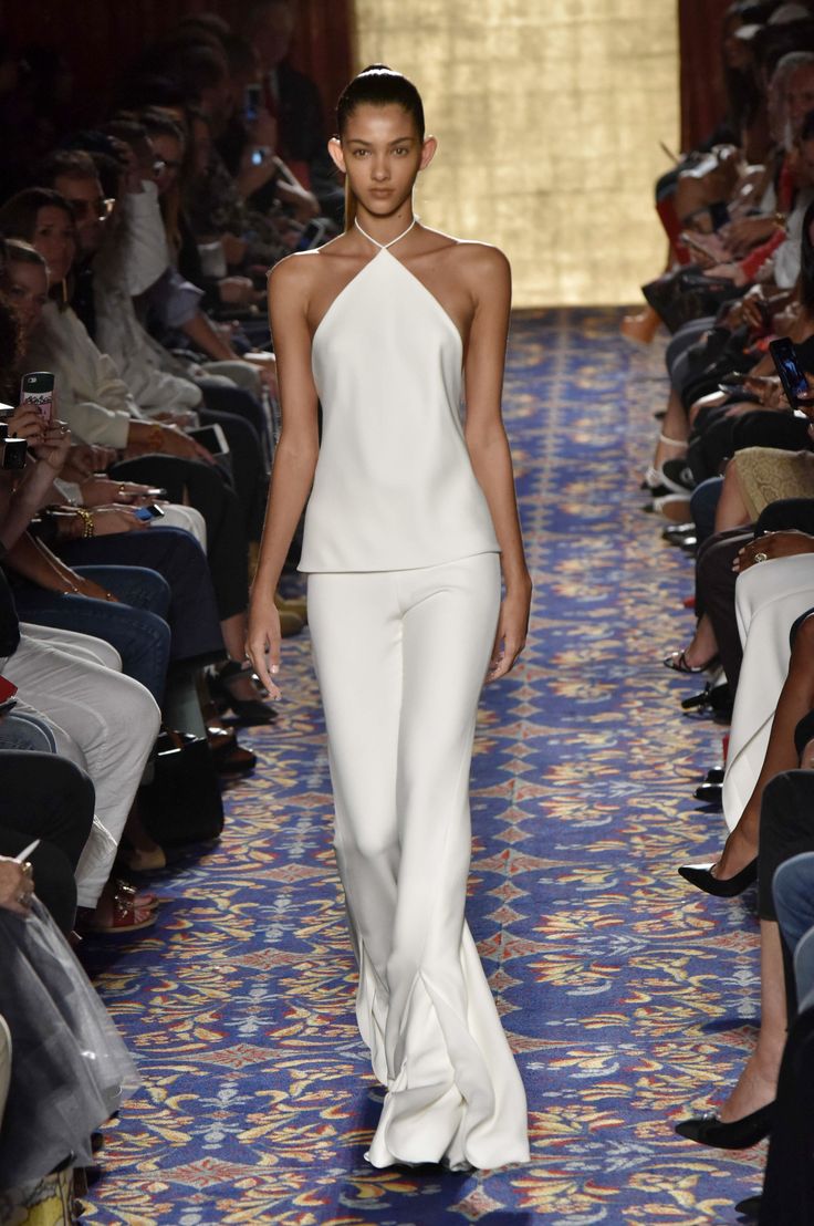 a fashion model walks down a runway in a white, sleek halter dress and flared pants. the dress features a halter neckline and the pants are widelegged and flowy. the model has a  minimalistic makeup look and her hair is pulled back in a sleek updo. the runway is covered in a carpet with blue and red patterns. there is an audience seated on either side of the runway, watching the model. the photo is taken from a slightly low angle, capturing the model from head to toe.