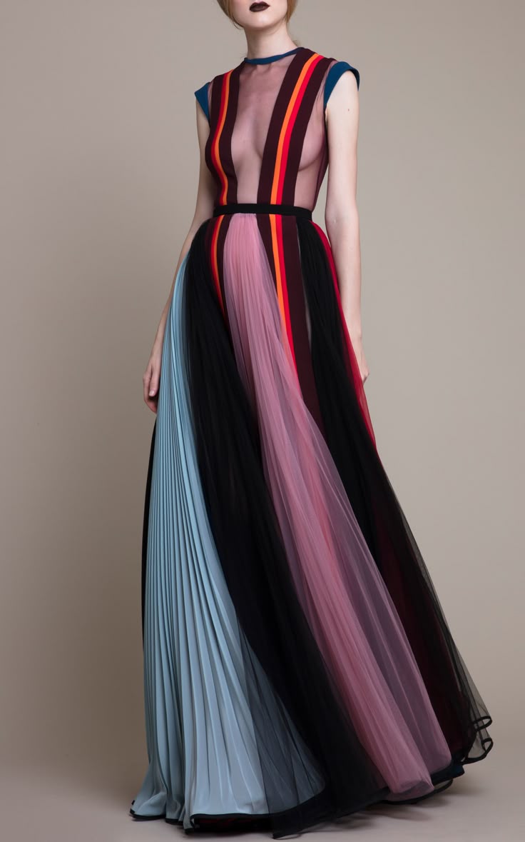 a woman stands wearing a floorlength gown with a sheer striped top and a pleated skirt in various colors. the top is sleeveless and has vertical stripes in red, orange, burgundy, and a muted blue. a black belt cinches the waist. the skirt consists of pleated sections in light blue, black tulle, and pink tulle. the woman has dark lipstick and is posing in a studio setting with a neutral backdrop.