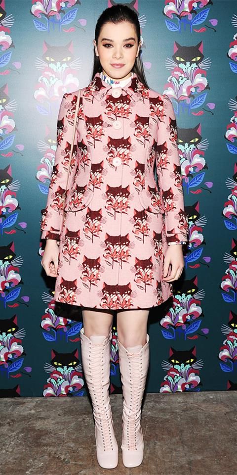 hailee steinfeld poses for a full shot portrait in front of a cat and floral patterned backdrop. she is wearing a pink coat dress with a matching print, along with light pink laceup kneehigh boots. she is looking at the camera.