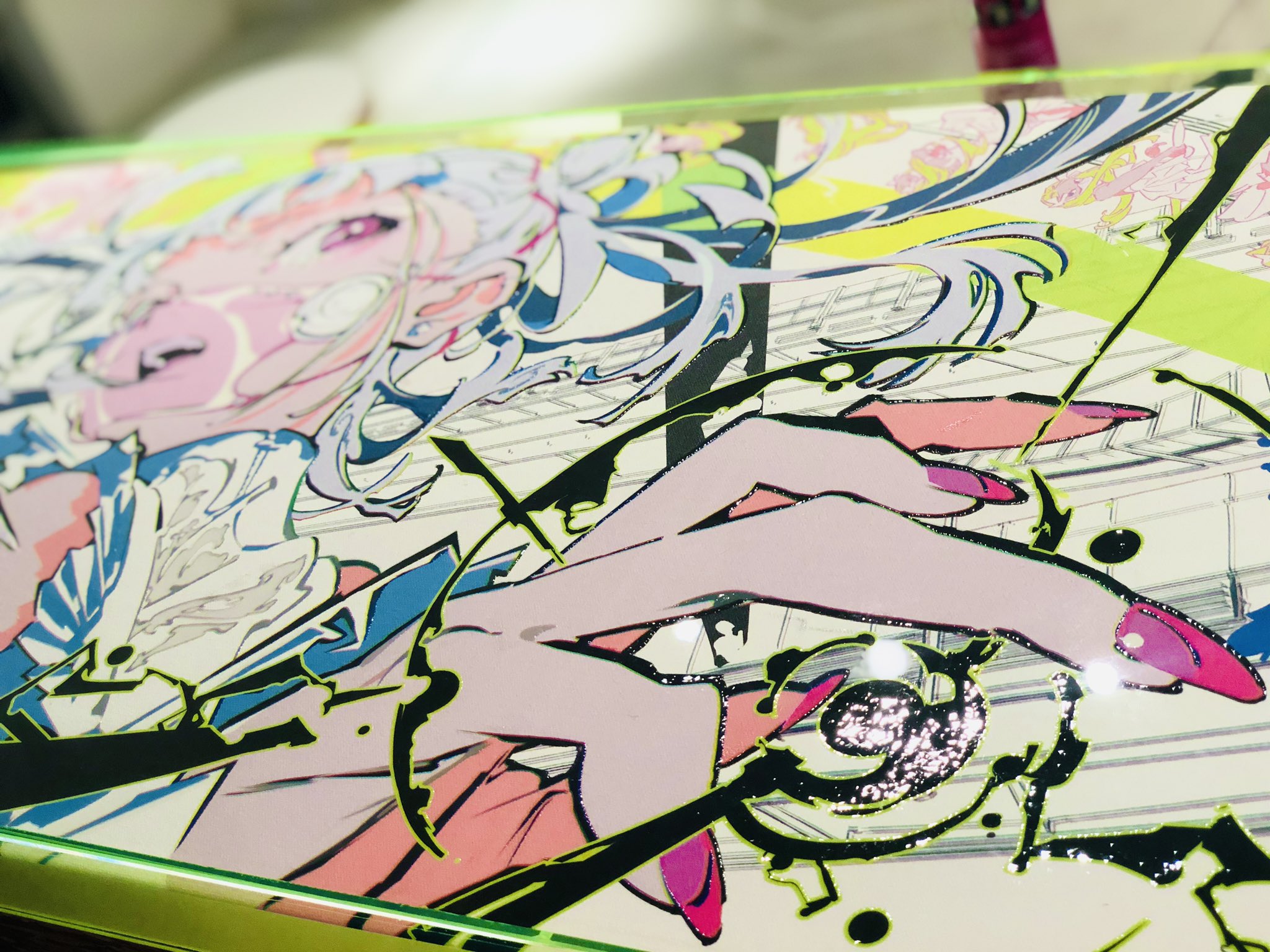 this image shows a closeup photograph of a colorful, graphic design piece featuring a stylized female character in an anime or manga style. the art is vibrant, with bold lines, abstract shapes, and a mix of pink, green, black, and white. it appears to be printed or painted on a surface with a glossy, reflective finish, possibly acrylic or resin. the closeup perspective emphasizes the details and textures of the artwork.
