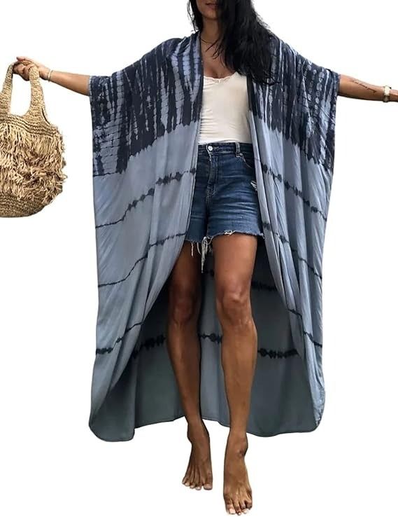 a woman is standing barefoot in a white top and denim shorts, wearing a blue and gray tiedye kimono. she is holding a beige fringe bag.