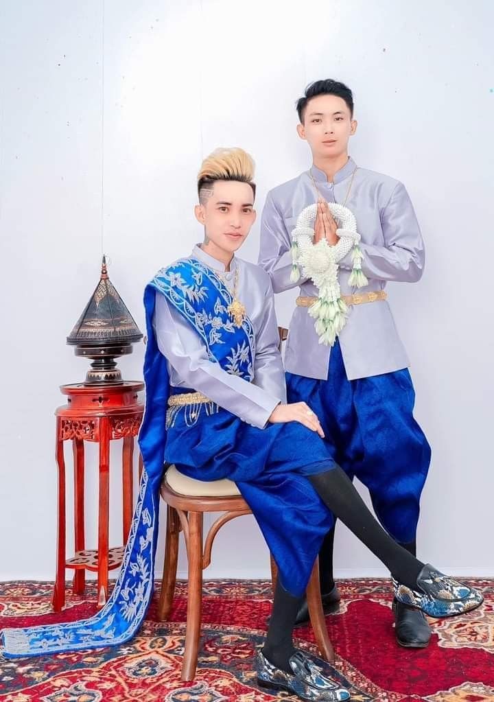 two men in traditional thai wedding attire, one seated on a chair and the other standing beside him, are smiling and posing for a photo. the man seated on the chair is wearing a silver top with blue and gold accents, blue pants, and black stockings with patterned loafers. the man standing is wearing a silver top with blue pants, black stockings and black loafers with a snake skin pattern. both men are wearing white garlands around their necks and are holding their hands in a praying position.
