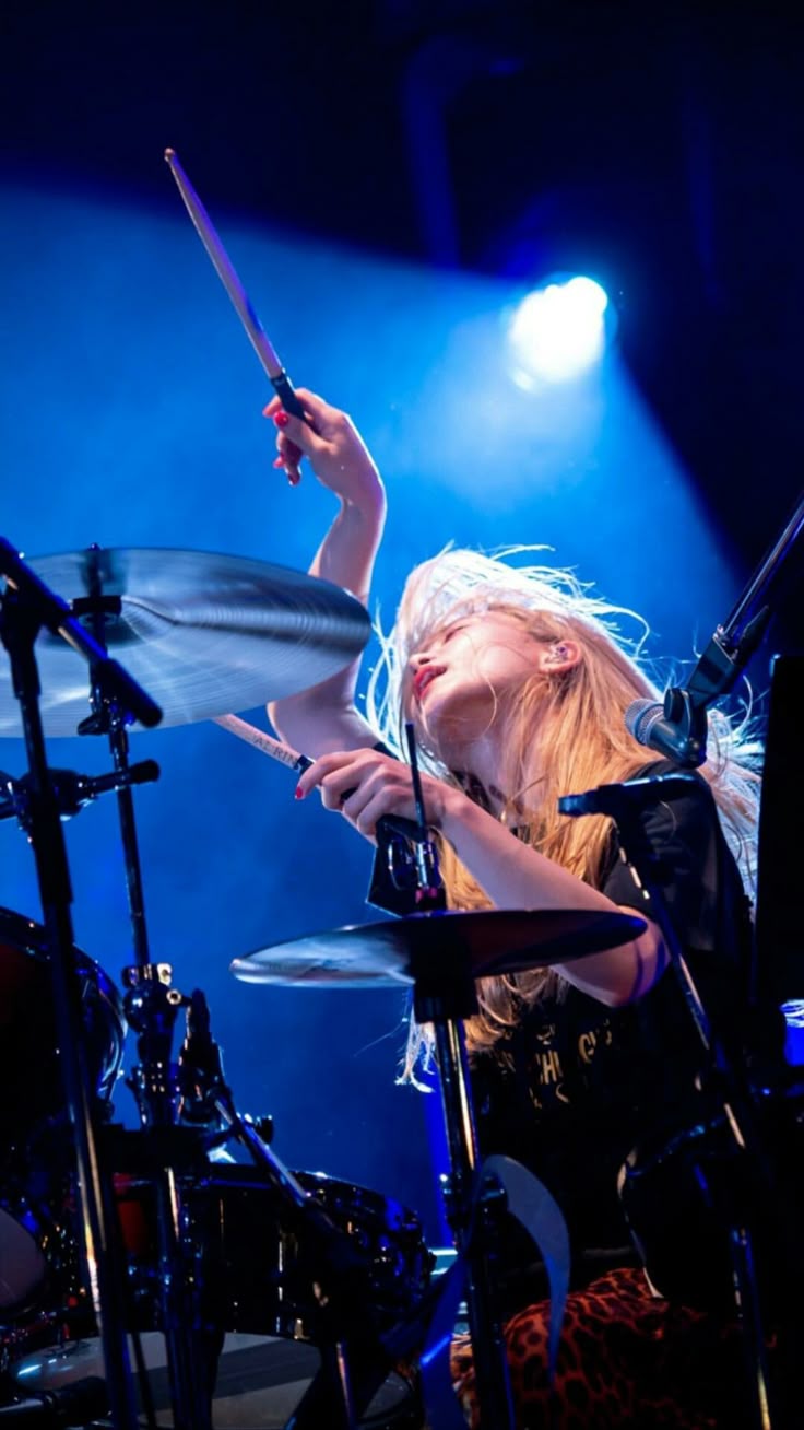 a woman with blonde hair is energetically playing the drums on a stage lit with blue lighting. she is holding drumsticks and striking the cymbals with force. the drum kit is visible, and she appears to be performing in a concert setting.