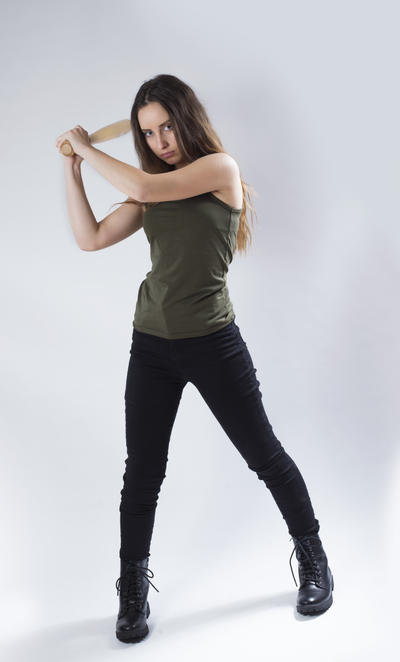 a fullbody shot of a young woman with long brown hair, wearing a green tank top and black pants and boots, standing and holding a baseball bat in a fierce, aggressive pose. she is looking directly at the camera with a serious expression. the background is plain gray, and the lighting is even. the overall mood is one of defiance and aggression.