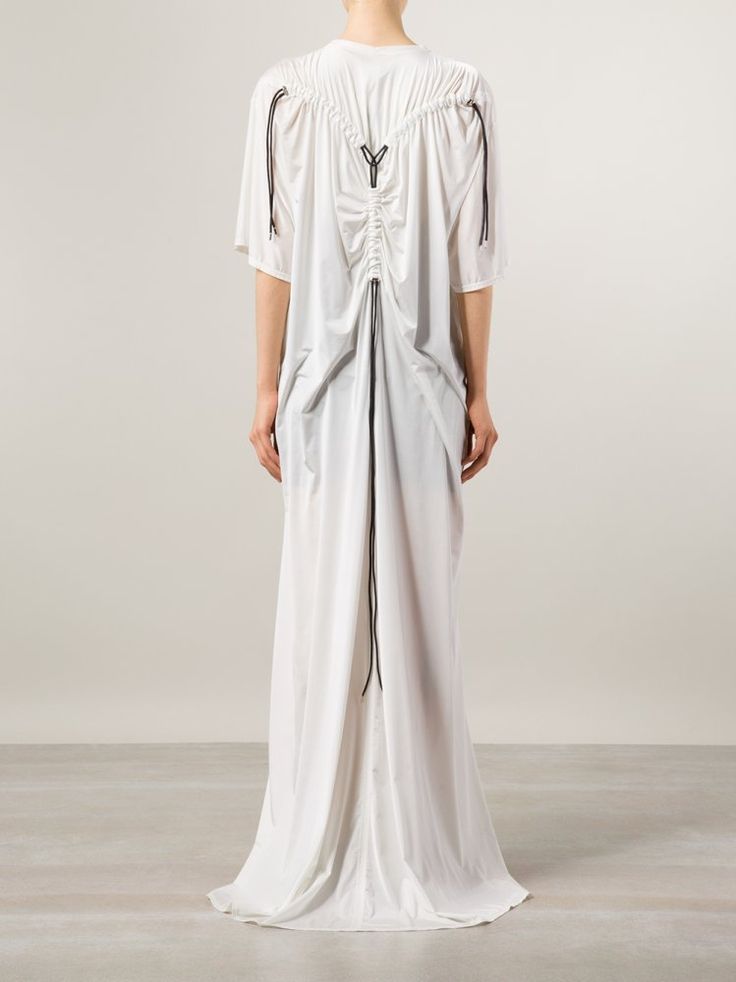 a fullbody, back view of a fairskinned person wearing a long white dress on a light beige floor. the floor continues into a light beige wall. the dress has short sleeves and ends in a floorlength train. a cinch detail on the back of the dress is laced with black drawstrings.