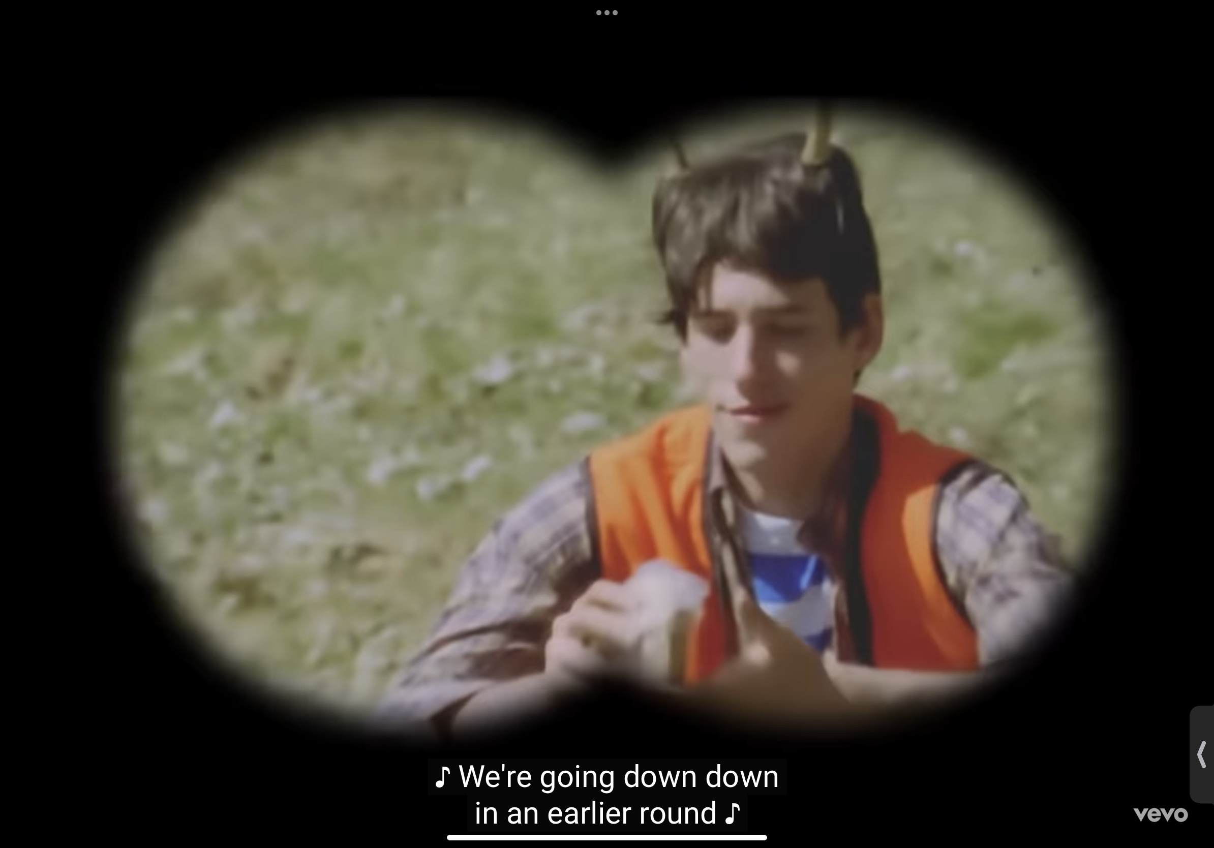 a young man wearing an orange vest and a striped shirt looks at the camera through binoculars. he is in an outdoor setting, surrounded by green grass. lyrics of a song are written on the bottom of the image. the word "vevo" is also at the bottom right of the image.