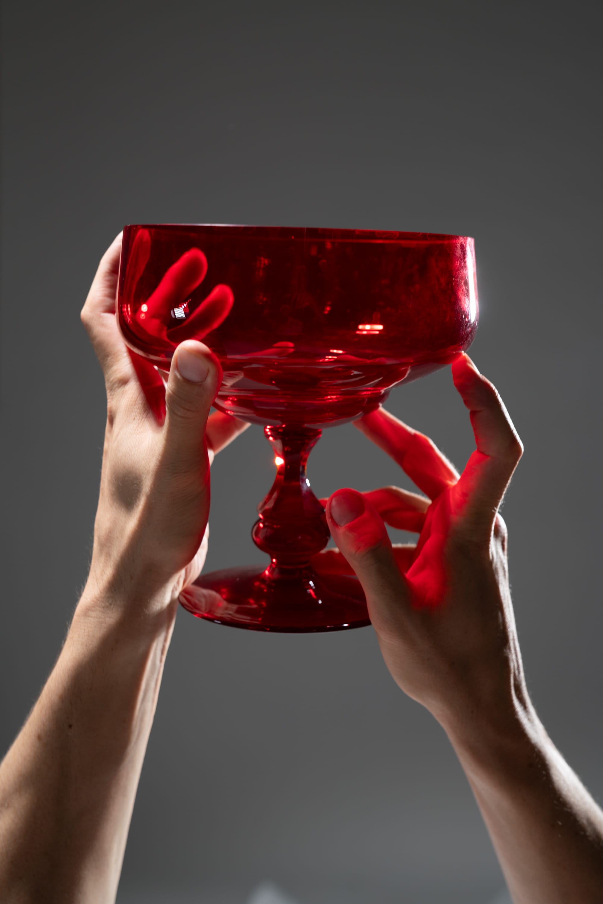 a pair of hands gracefully holds up a vibrant red glass against a dark gray background, illuminated by soft studio lighting. the transparency of the glass allows light to pass through, casting a subtle glow on the hands and highlighting the intricate details of its design. the image evokes a sense of elegance and fragility.