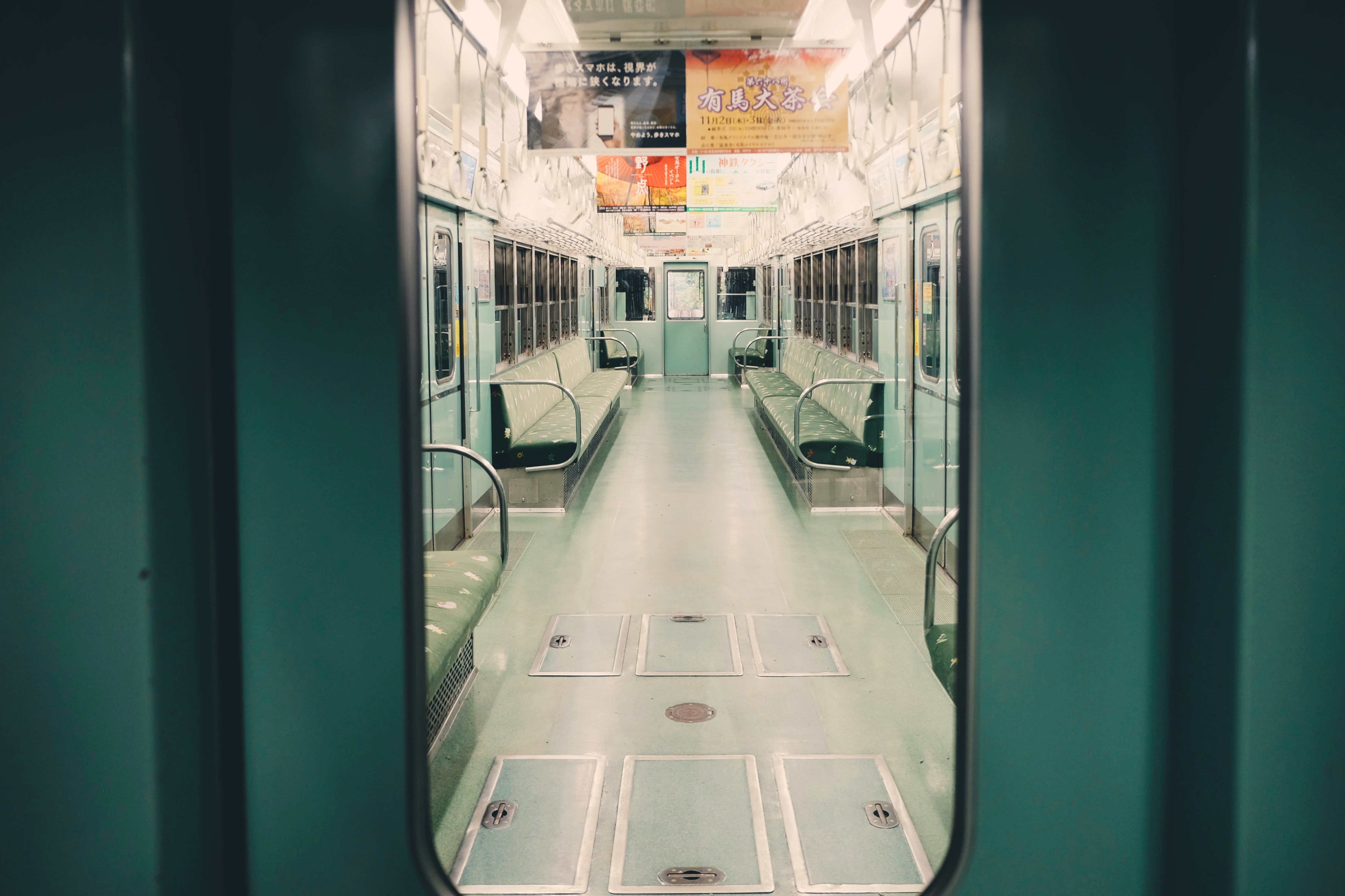 a view of an empty subway car through an open doorway. the subway car features green seats, metal handles, and advertisements on the walls above the seats. the floor has rectangular access panels. the lighting is soft and even, illuminating the interior of the train car. the doorway provides a framed view of the symmetrical layout of the subway car.