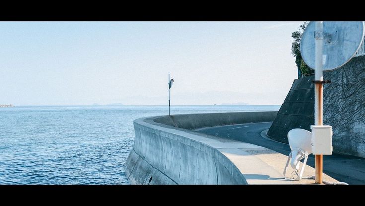 the image depicts a tranquil coastal scene. a curved road winds along a high concrete seawall that juts out into a calm, blue ocean. the sky is clear and bright. in the background, a faint horizon line can be seen.  a pole with a small antenna or satellite dish stands near the wall, and another, larger dishlike antenna is situated lower down on the wall near the road. the overall feeling is serene and peaceful. the scene appears to be in japan, judging by the style of the infrastructure.