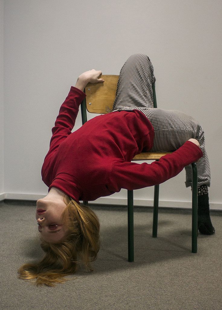 a young woman is sitting upside down on a chair. she is wearing a red sweater and black and white checkered pants. her long brown hair is cascading down to the floor. she is looking straight ahead with a serious expression on her face.