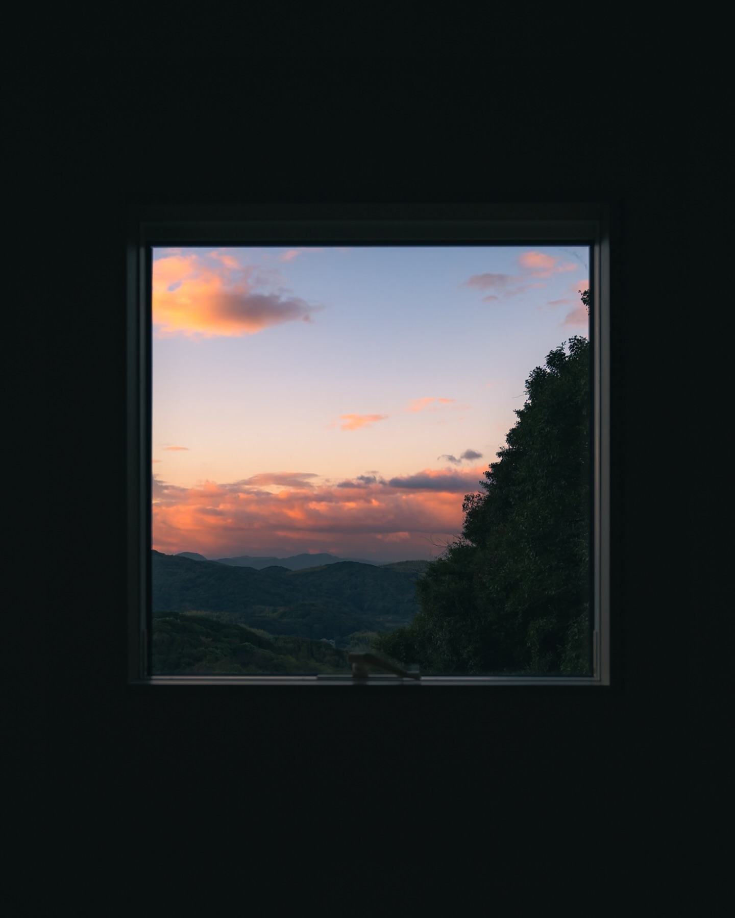 the image shows a square window in a dark room.  the window provides a stunning view of a sunset over a rolling landscape of hills and mountains.  the sky is a mix of soft pinks and oranges, contrasting against the darker silhouette of the trees and distant hills. the scene is peaceful and serene.  the darkness of the room emphasizes the vibrant colors of the sunset outside.
