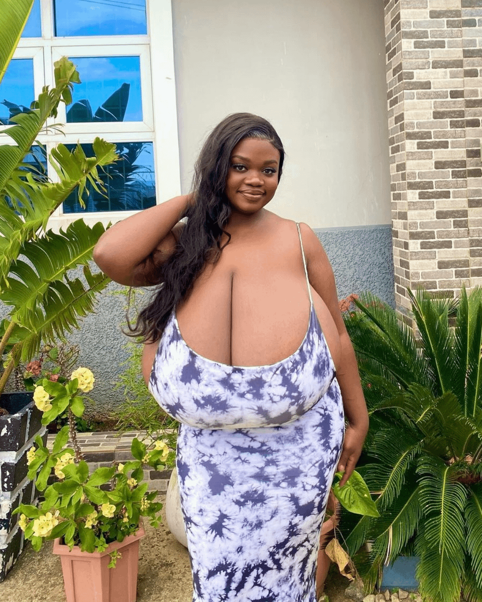 the image shows a darkskinned woman with very large breasts wearing a long, formfitting tiedye maxi dress in shades of blue and white. she has long black hair and is smiling confidently at the camera while standing in a garden setting in front of a house with a brick wall.  the background includes lush green plants and a window. the overall impression is one of body positivity and selfassuredness.
