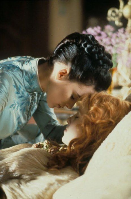 a woman in a blue dress, with her hair tied back, is looking down at another woman lying in bed.  the women look to be of the victorian era, and the scene could be from a movie or a play. the scene is warm and caring, and the women seem to have a close relationship.  the scene is set in a room with a large bed.